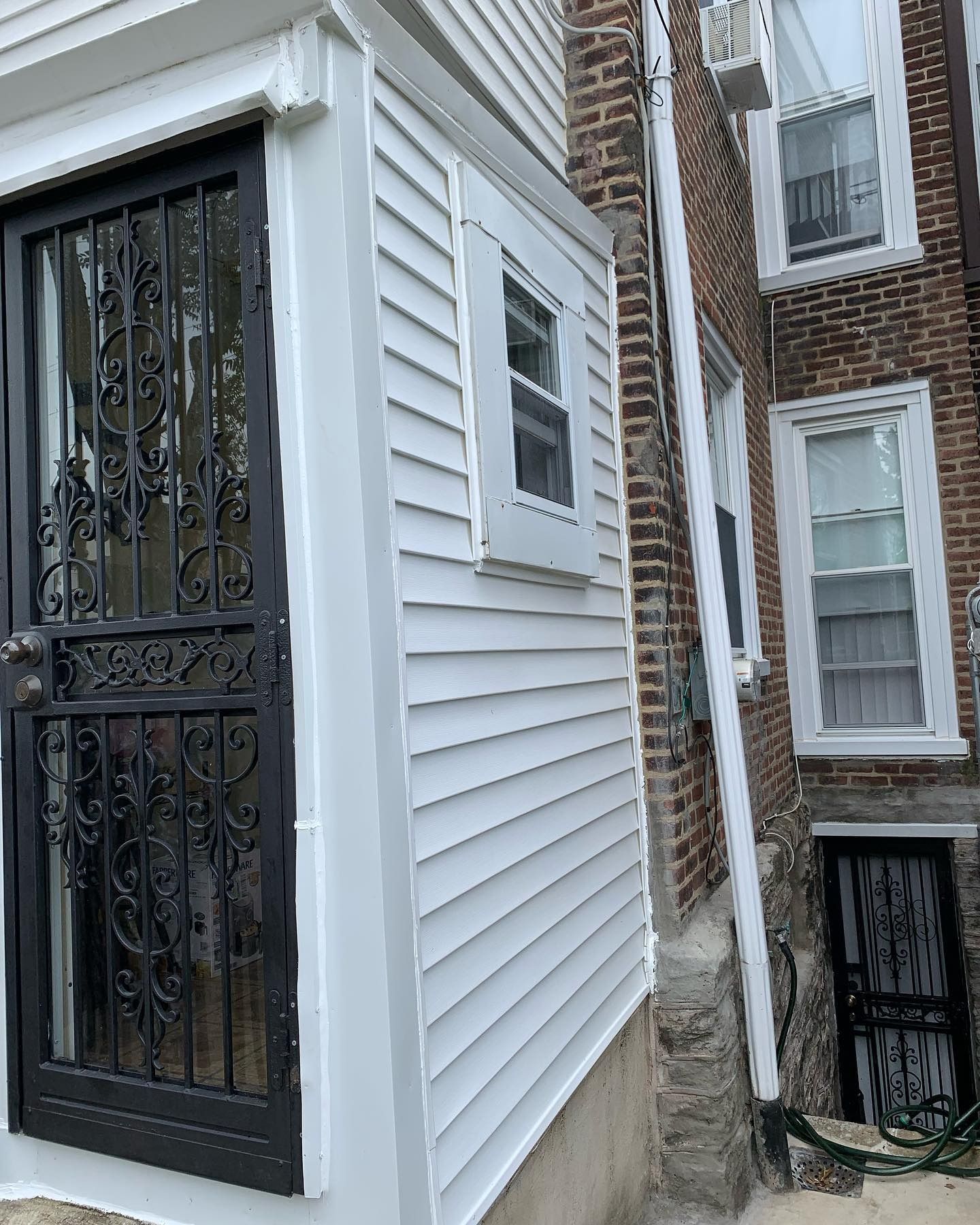 Narrow alley beside a white-sided building with a black security door and windows, next to a brick house.