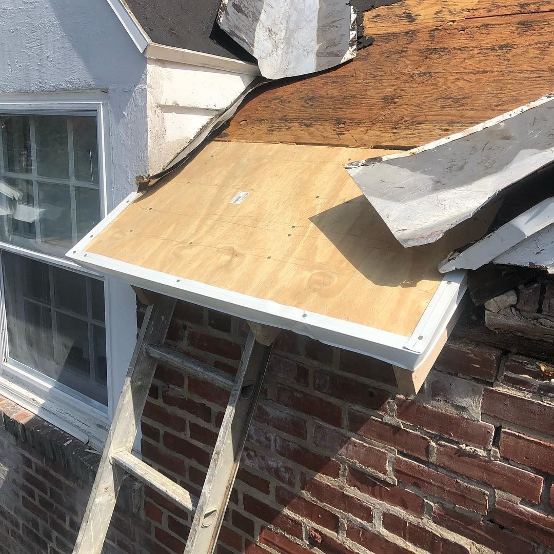 Damaged roof with exposed plywood and broken shingles above a brick wall and window