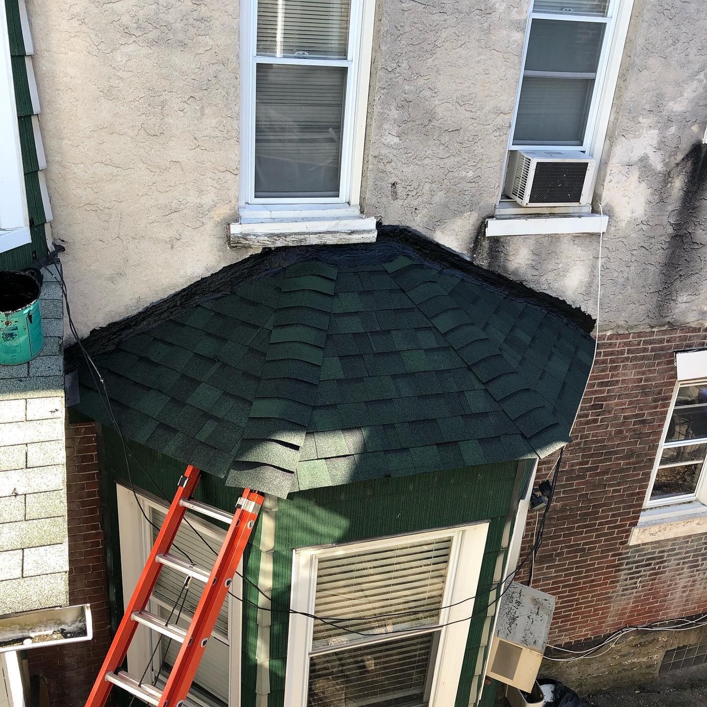 Red ladder leaning against a green bay window under a dark shingled roof on a brick building