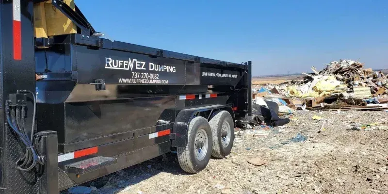 A black dump trailer parked on a dirt lot next to a large pile of debris under a clear blue sky. Dumpster service Pflugerville TX
