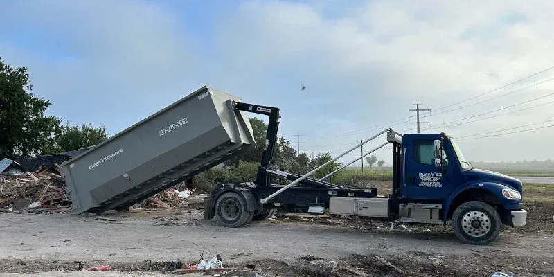 A blue roll-off dump truck in an outdoor setting, actively lowering a large grey metal dumpster onto a gravel lot. A blue roll-off dump truck in an outdoor setting, actively lowering a large grey metal dumpster onto a gravel lot.