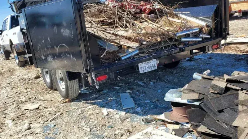 A black dump trailer filled with debris and tree branches sits on a dirt lot, with a pile of shingles in the foreground. Construction dumpster rentals Pflugerville TX