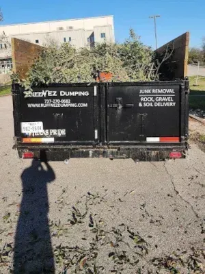 A black dump trailer filled with tree trimmings parked outdoors, displaying contact information for a junk removal service. Local dumpster Pflugerville TX