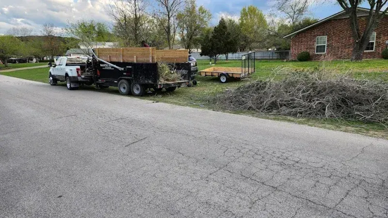 A white truck towing a black dump trailer filled with yard waste, with a smaller flatbed trailer parked on a lawn nearby. Local dumpsters for rent in Pflugerville TX