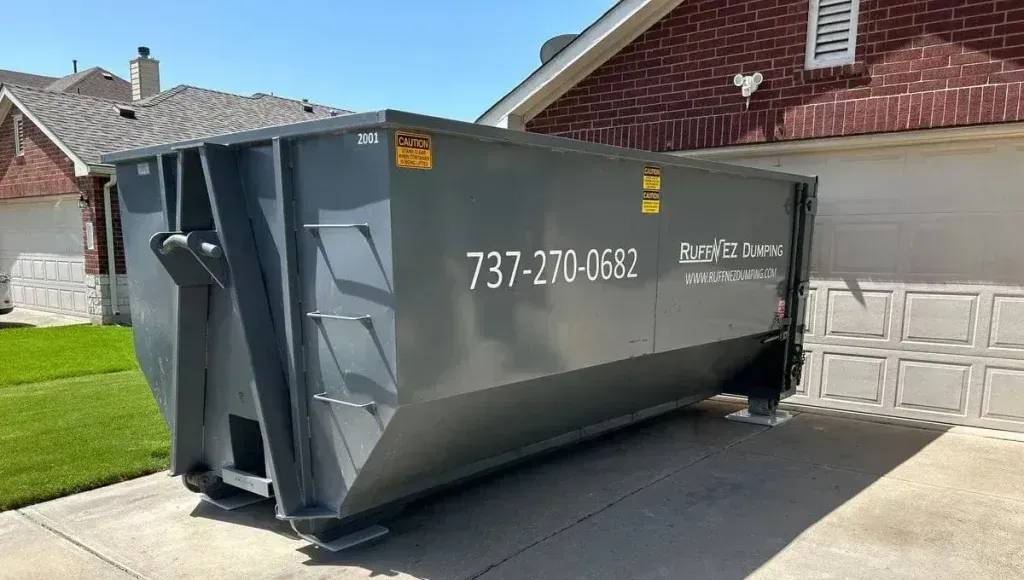 A large, grey metal dumpster sits in a residential driveway in front of a house and garage. Rent a dumpster in Pflugerville