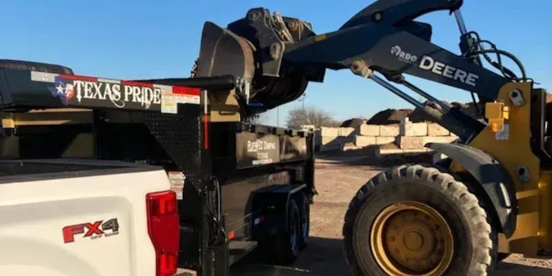 A yellow loader uses its bucket to lift the gooseneck hitch of a black Texas Pride trailer onto a pickup truck. Rent dumpster in Pflugerville