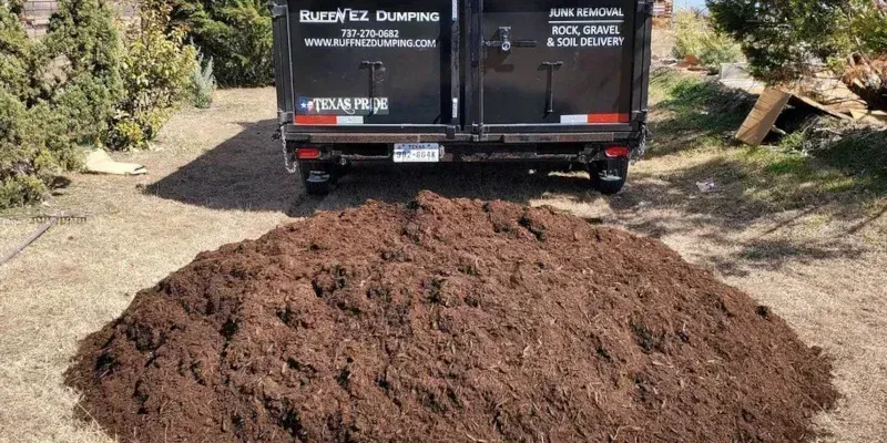 A large pile of dark brown mulch sitting on the ground in front of a black dump trailer in a yard. Tree mulch delivery in Pflugerville TX