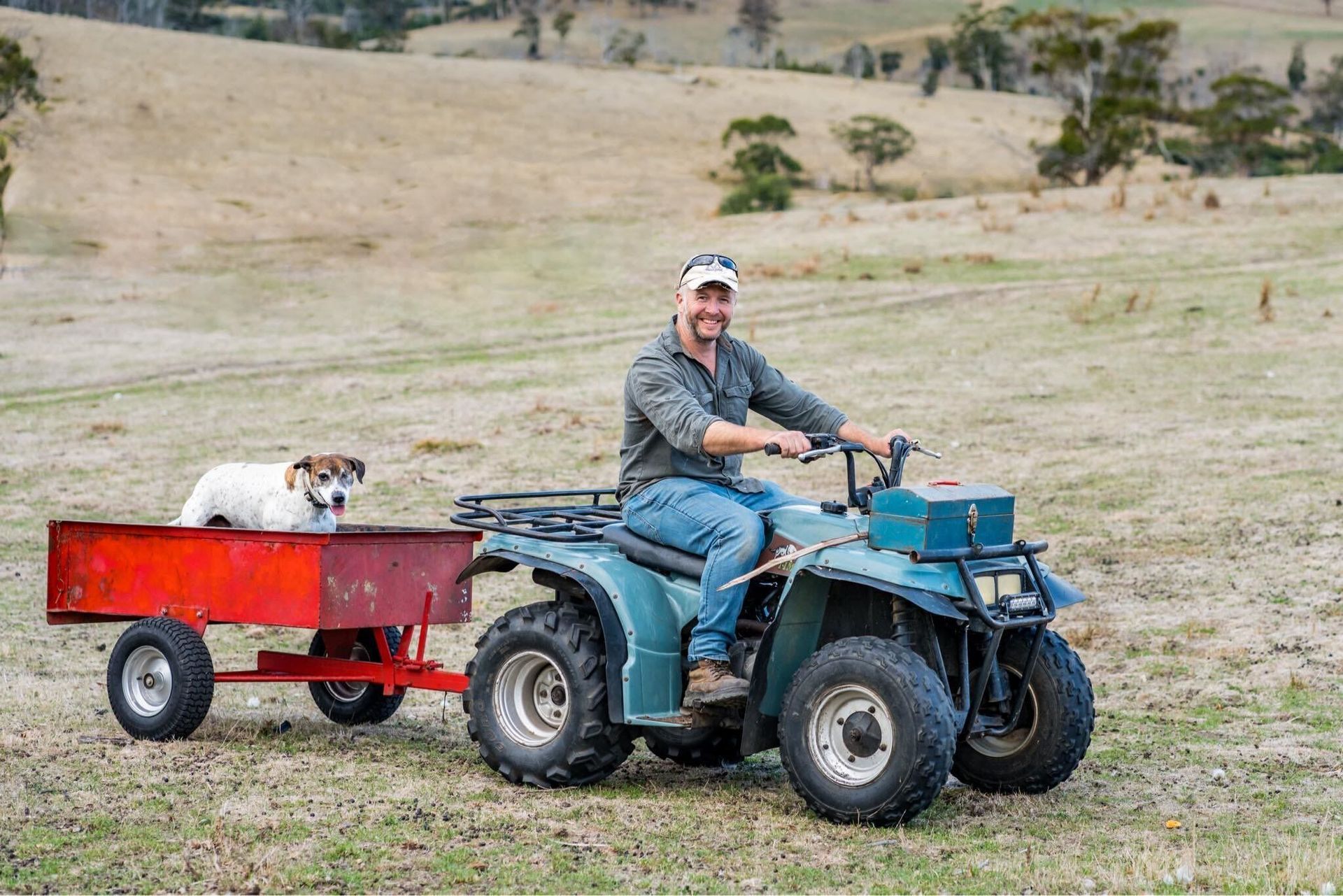 A man is riding a four wheeler with a dog in the trailer.