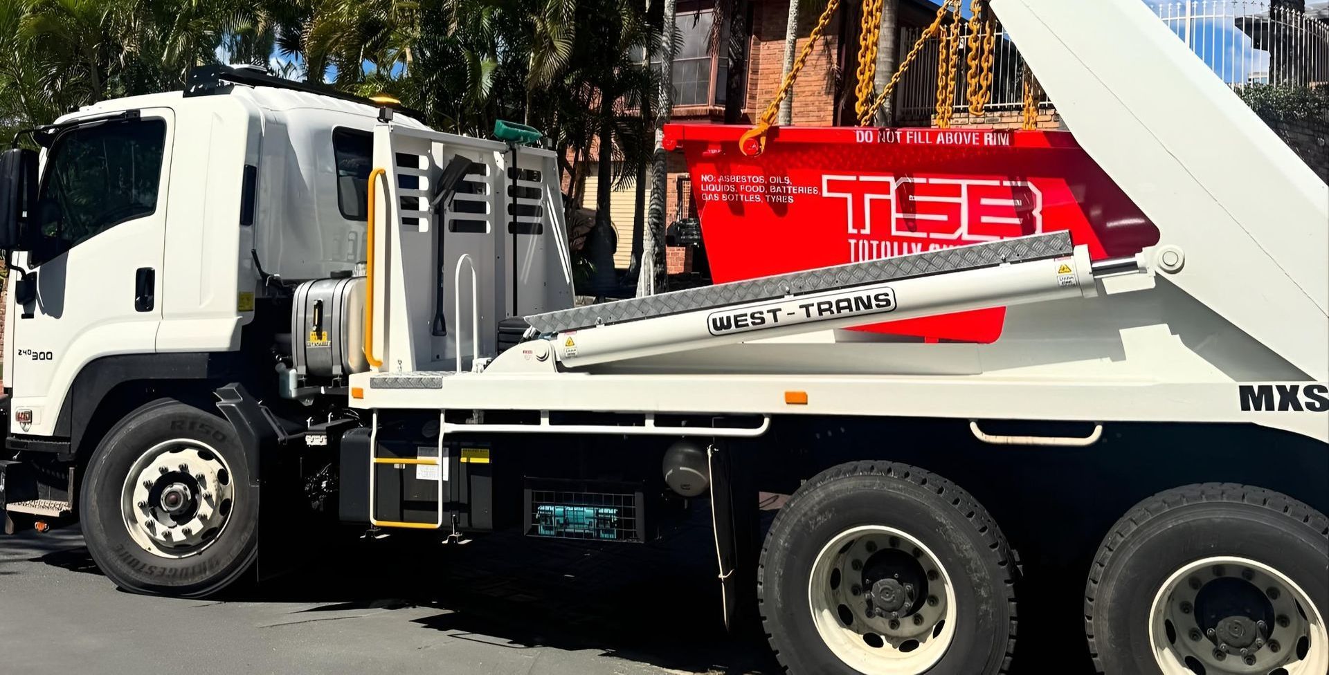 White skip bin truck lifting a bright red waste container labeled on a residential street