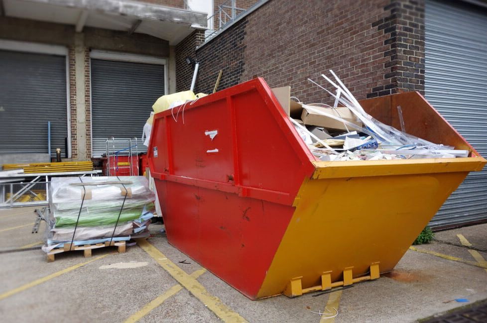 A Red and Yellow Dumpster is Sitting in Front of a Brick Building — Totally Skip Bins in Jimboomba, QLD
