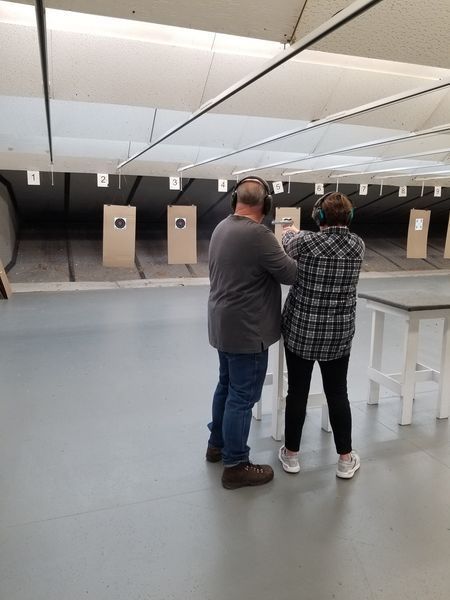 Man assisting woman at a shooting range; both wear ear protection, shooting towards targets.