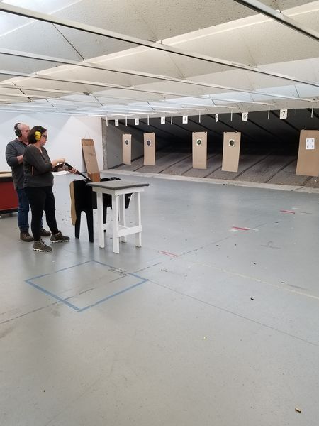 Two people at a shooting range, one coaching the other; targets in the distance, gray concrete floor.