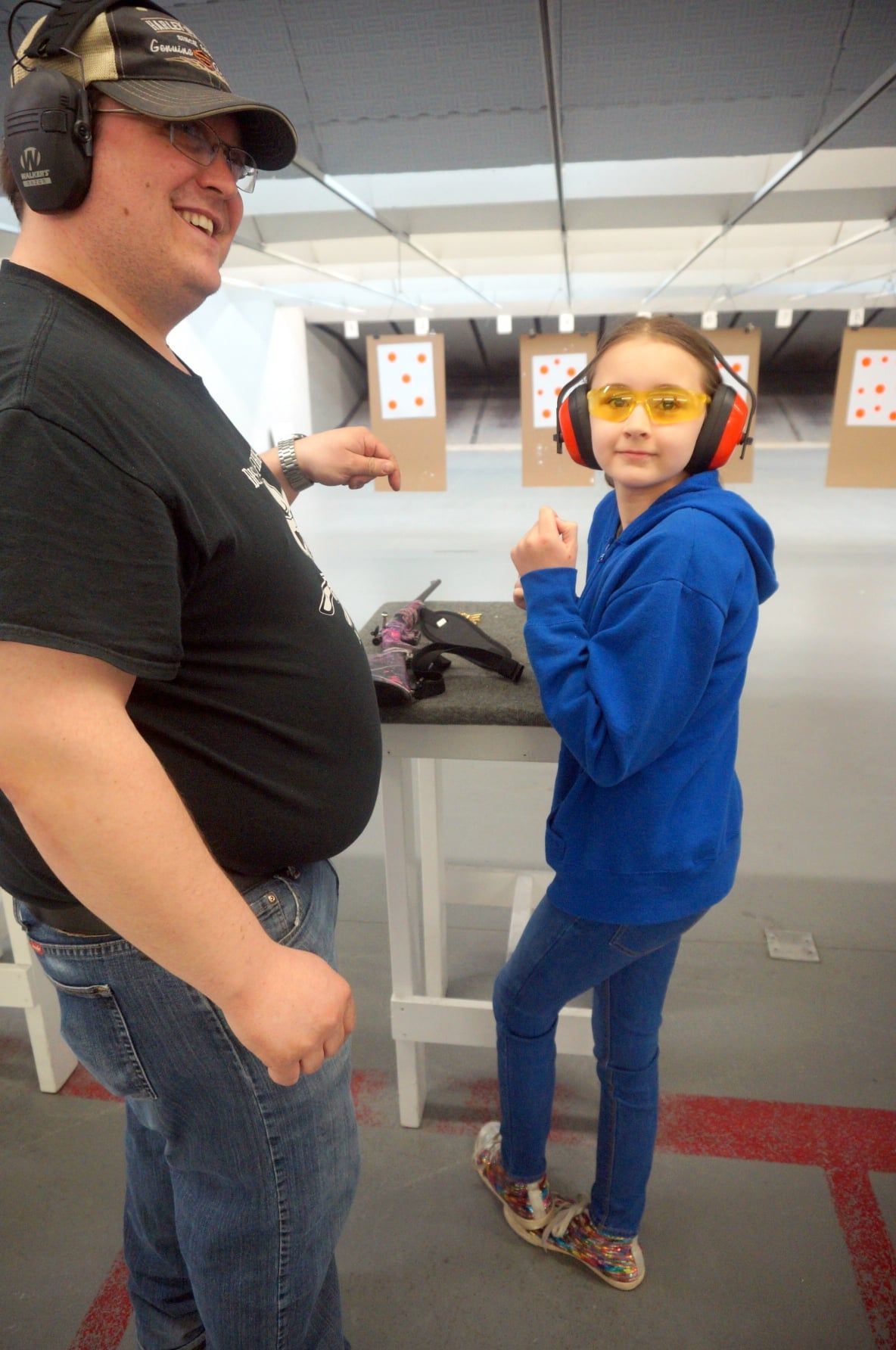 Man and young girl at a shooting range. Man points, girl smiles and wears safety glasses and earmuffs.