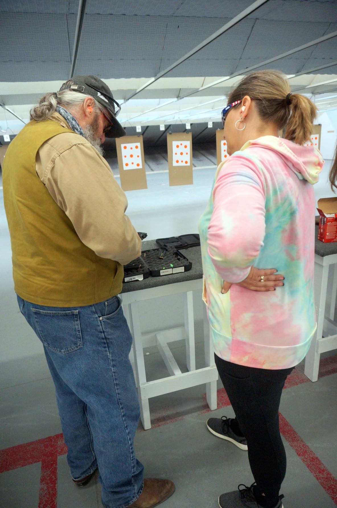 Man showing woman firearms at a shooting range; both are wearing glasses, looking at guns.