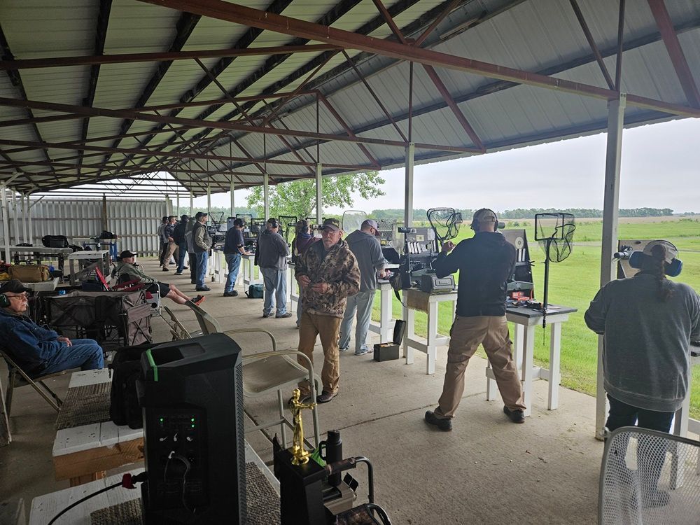 People shooting at a range. Under a covered area, they stand at tables, with targets in the distance, and a grassy field.