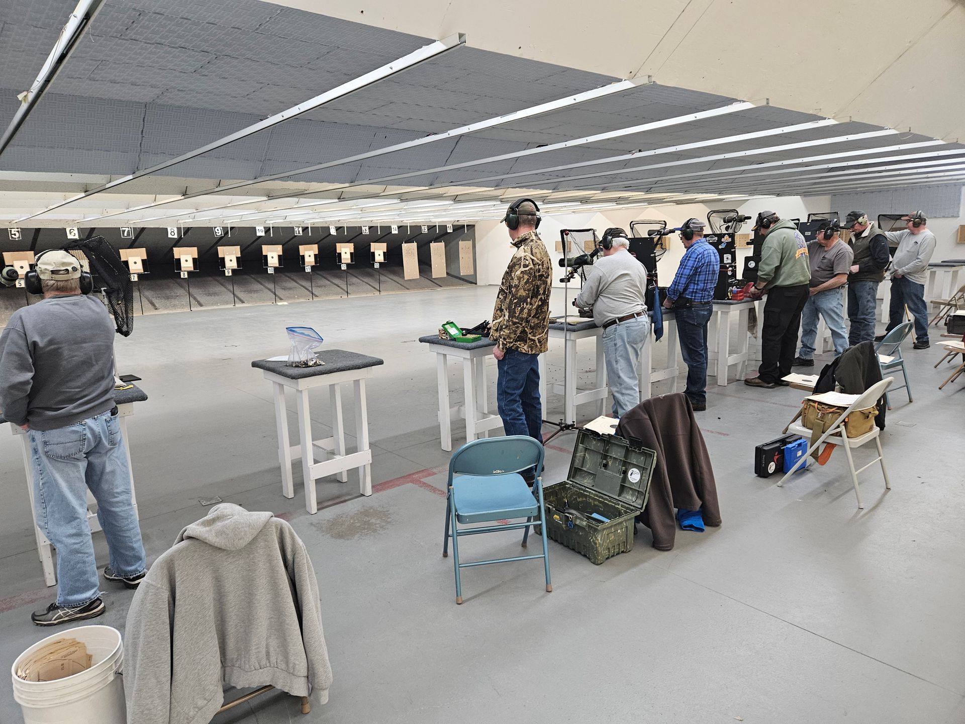 People at a shooting range, aiming and handling firearms. Indoor range, gray walls, white targets.