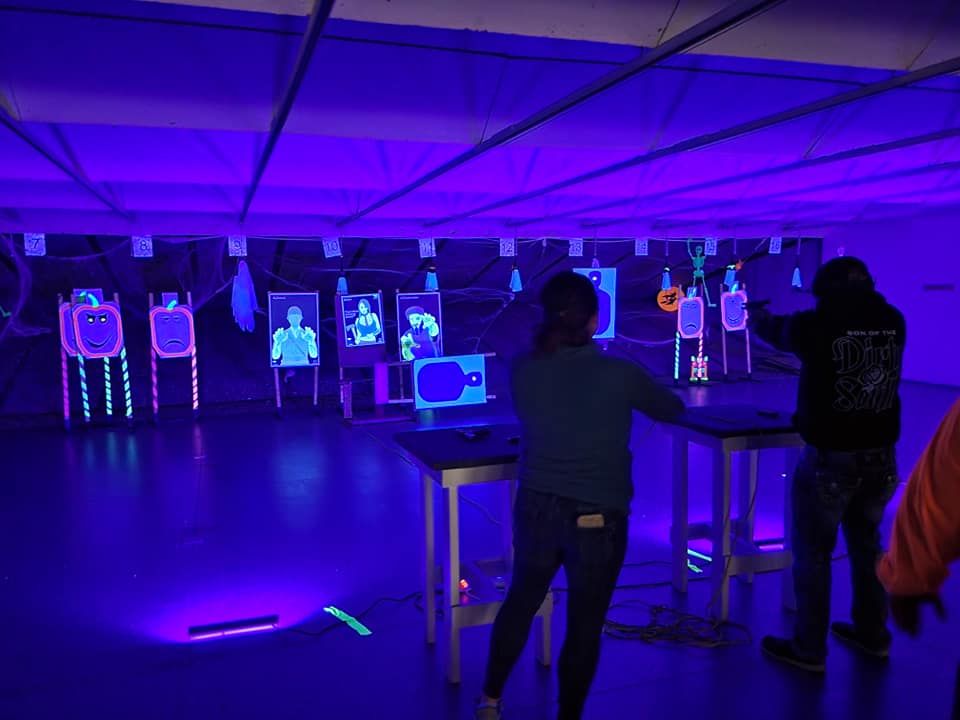 People shooting targets in a neon-lit gun range; purple light illuminates the scene.