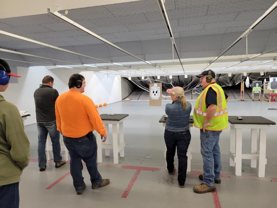 People at an indoor shooting range. One woman aims a handgun, supervised by two men. Target at distance.