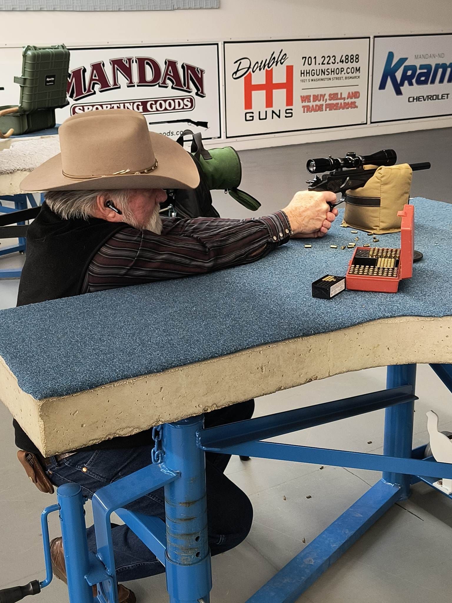 Man wearing a cowboy hat aiming a rifle at a shooting range. He’s wearing ear protection and is seated at a bench.