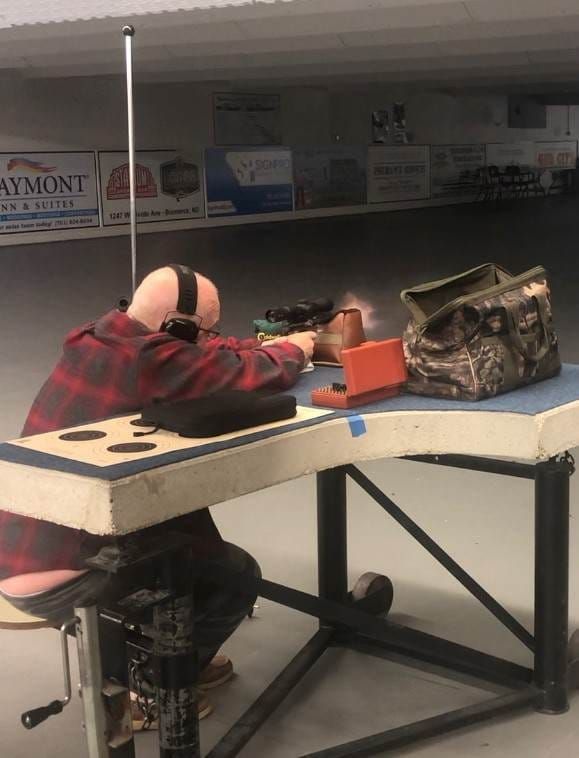 Person in a red plaid shirt aiming a rifle at a shooting range. He is seated, wearing earmuffs.