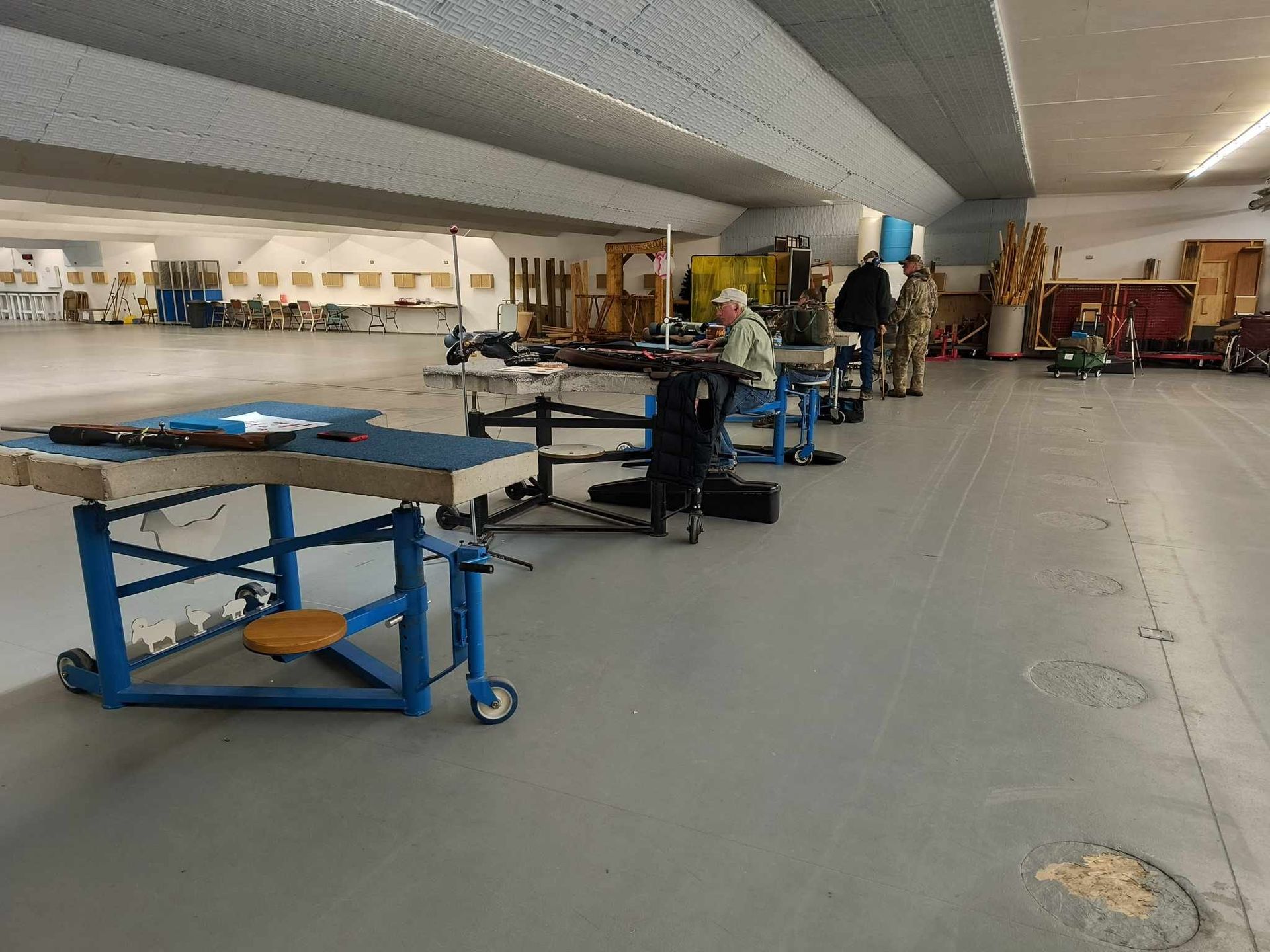 Indoor shooting range with people and firearms at tables under a long, light-colored ceiling.