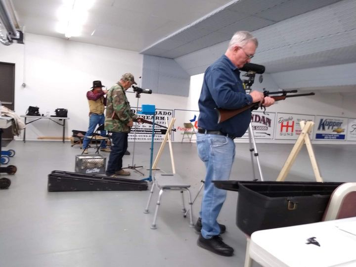 Men at indoor gun range, aiming rifles at targets.