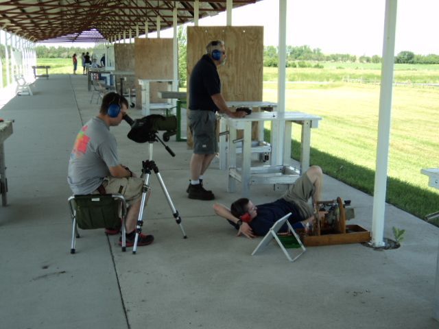People at a shooting range, using firearms. One person is lying down, others standing or sitting. Outdoors, sunny.