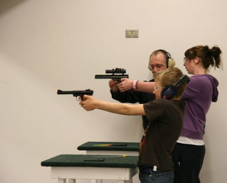 Three people at a shooting range: a man and two children, all aiming pistols. All wear ear protection.