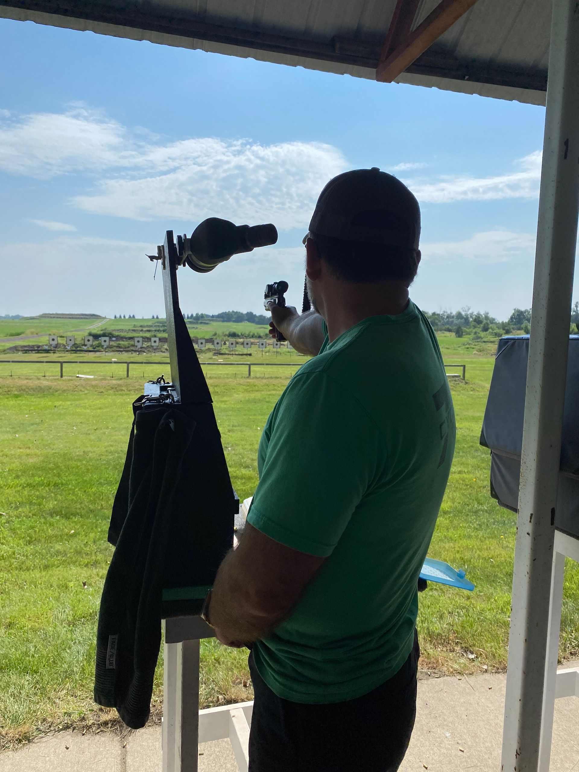 Man in green shirt shooting a rifle at an outdoor range on a sunny day.