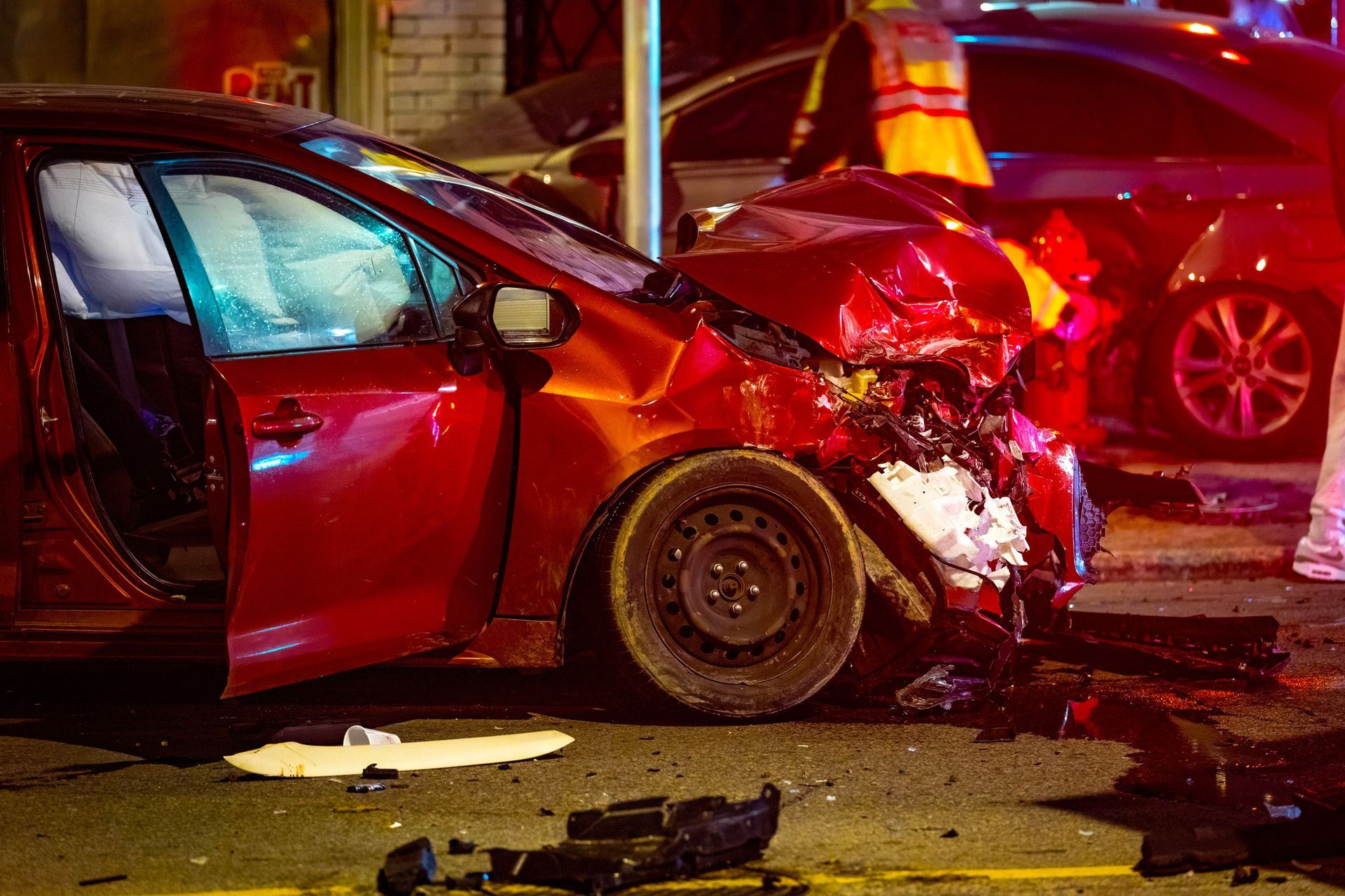 Person in a suit reviewing documents on a desk with a calculator, gavel, car, and credit cards. Red car, heavily damaged in a crash, deployed airbags, street scene.