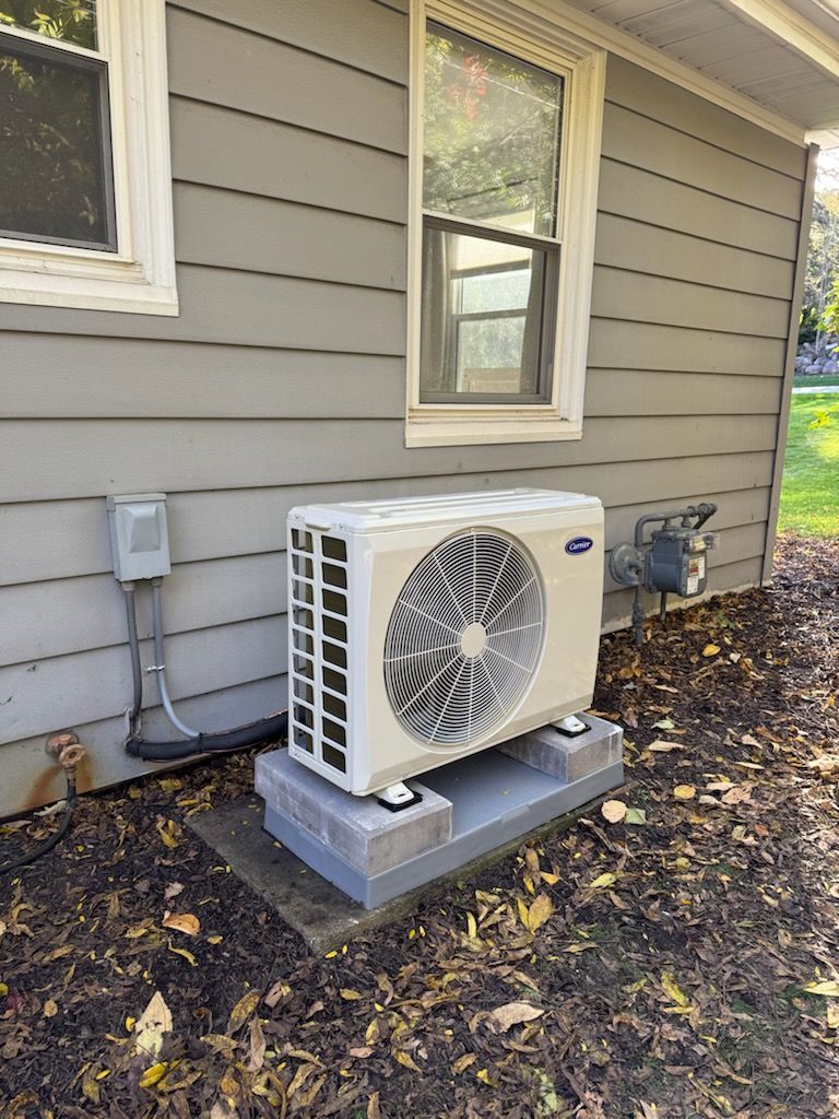 Outdoor HVAC unit beside a gray house wall on a concrete pad, with fallen leaves around it.