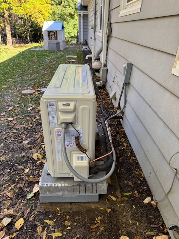 Outdoor HVAC unit beside a house, with hoses and wiring on a concrete pad in a yard.