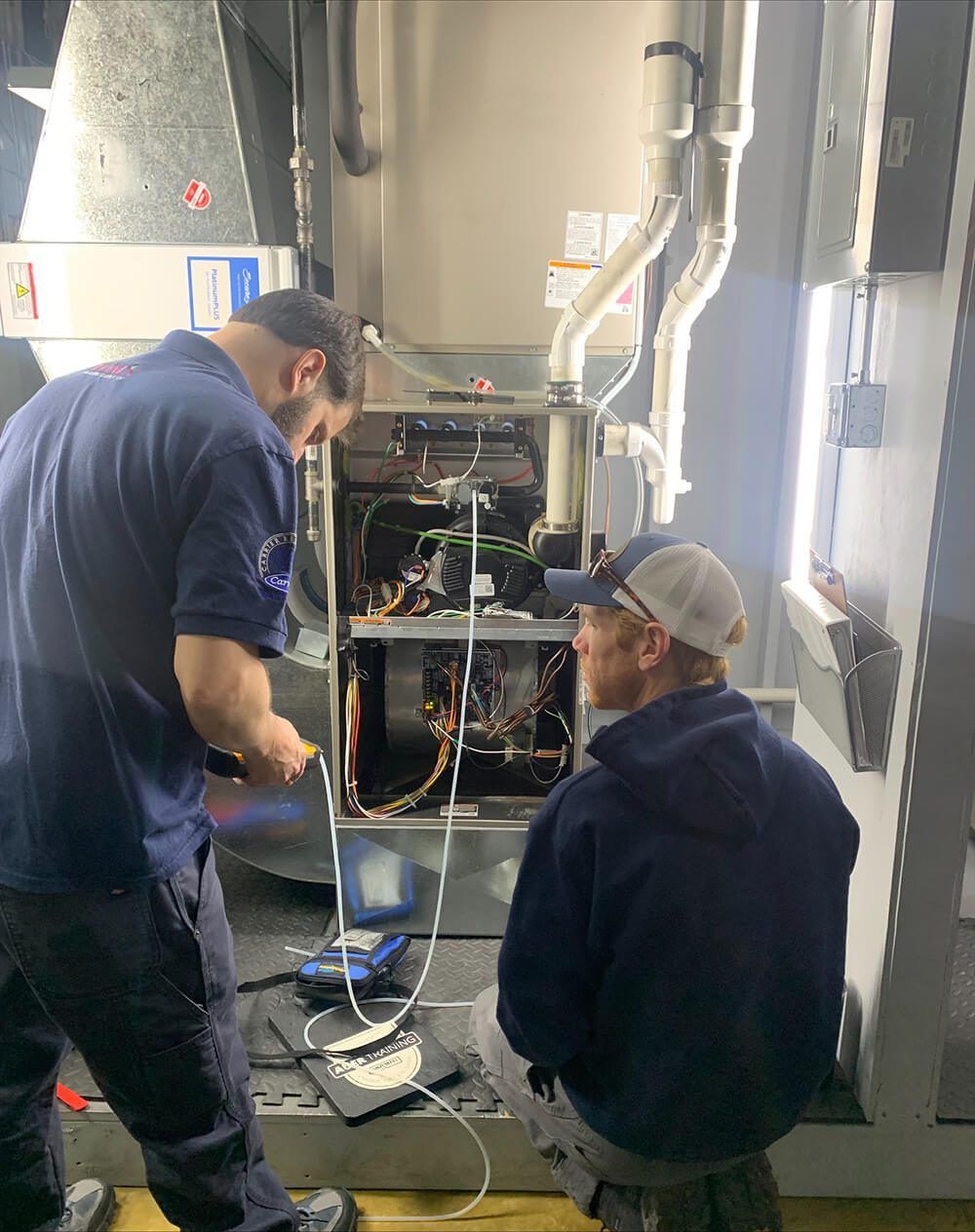 Two technicians inspect an open HVAC unit in a mechanical room, with ductwork and cables visible.