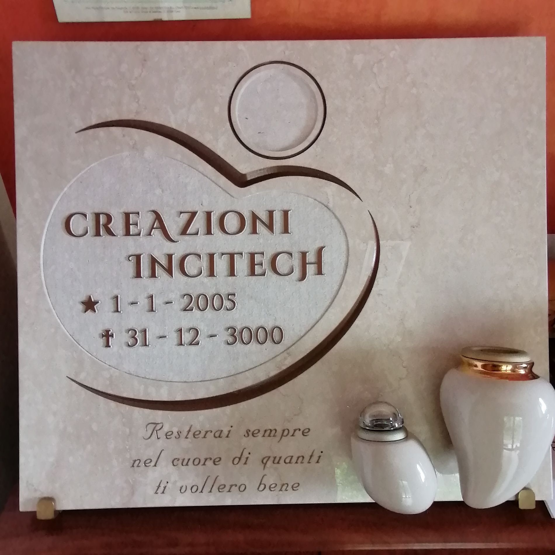 Targa commemorativa beige con incisa la scritta “CREAZIONI INTECCH” e le date, accanto a due piccole urne in ceramica.