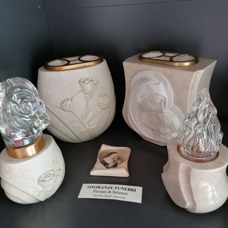 Candele commemorative decorative in ceramica con rilievi floreali e coperchi in vetro, esposte su uno scaffale scuro.