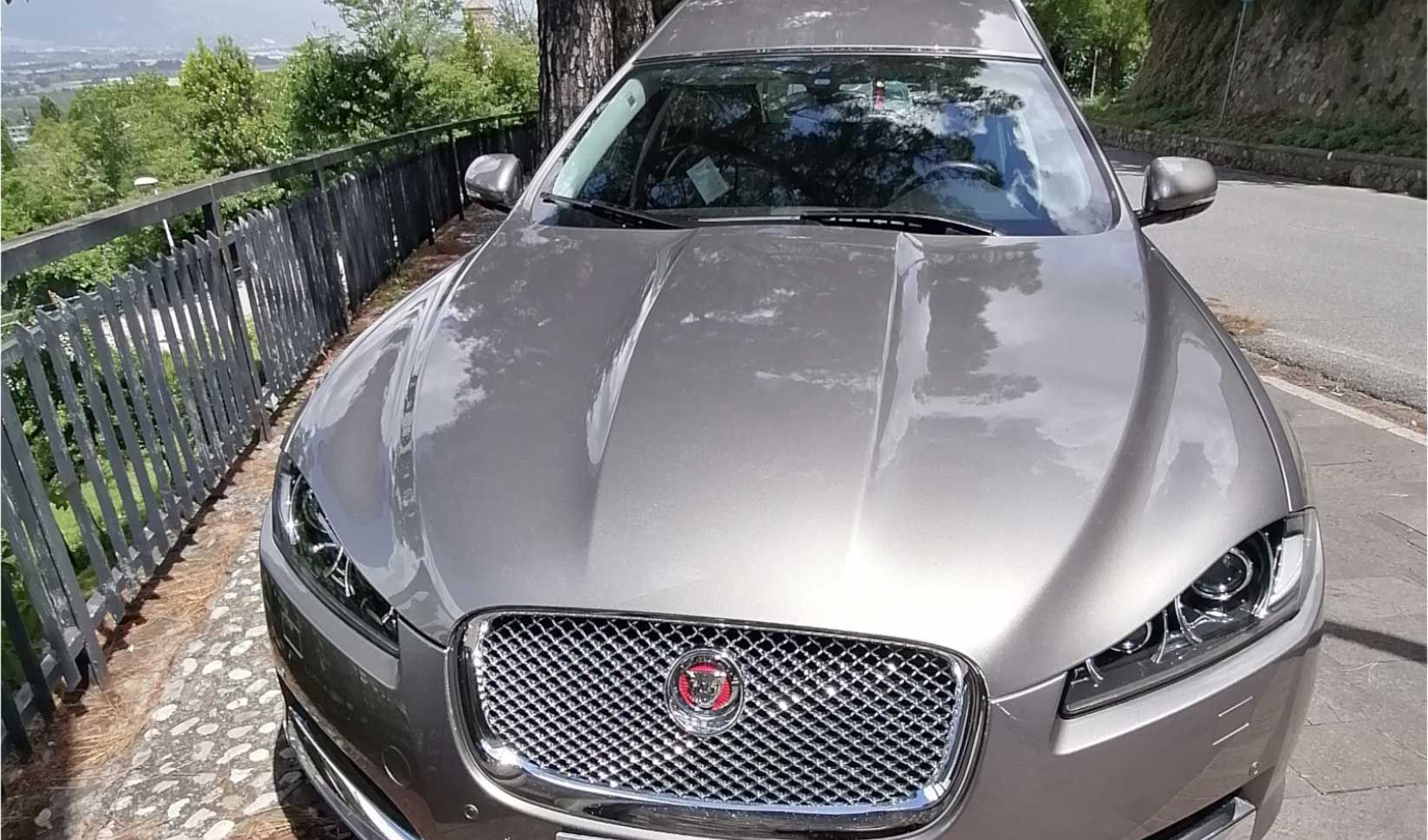 Vista frontale di una auto funebre Jaguar grigia parcheggiata sul ciglio della strada accanto a una recinzione metallica