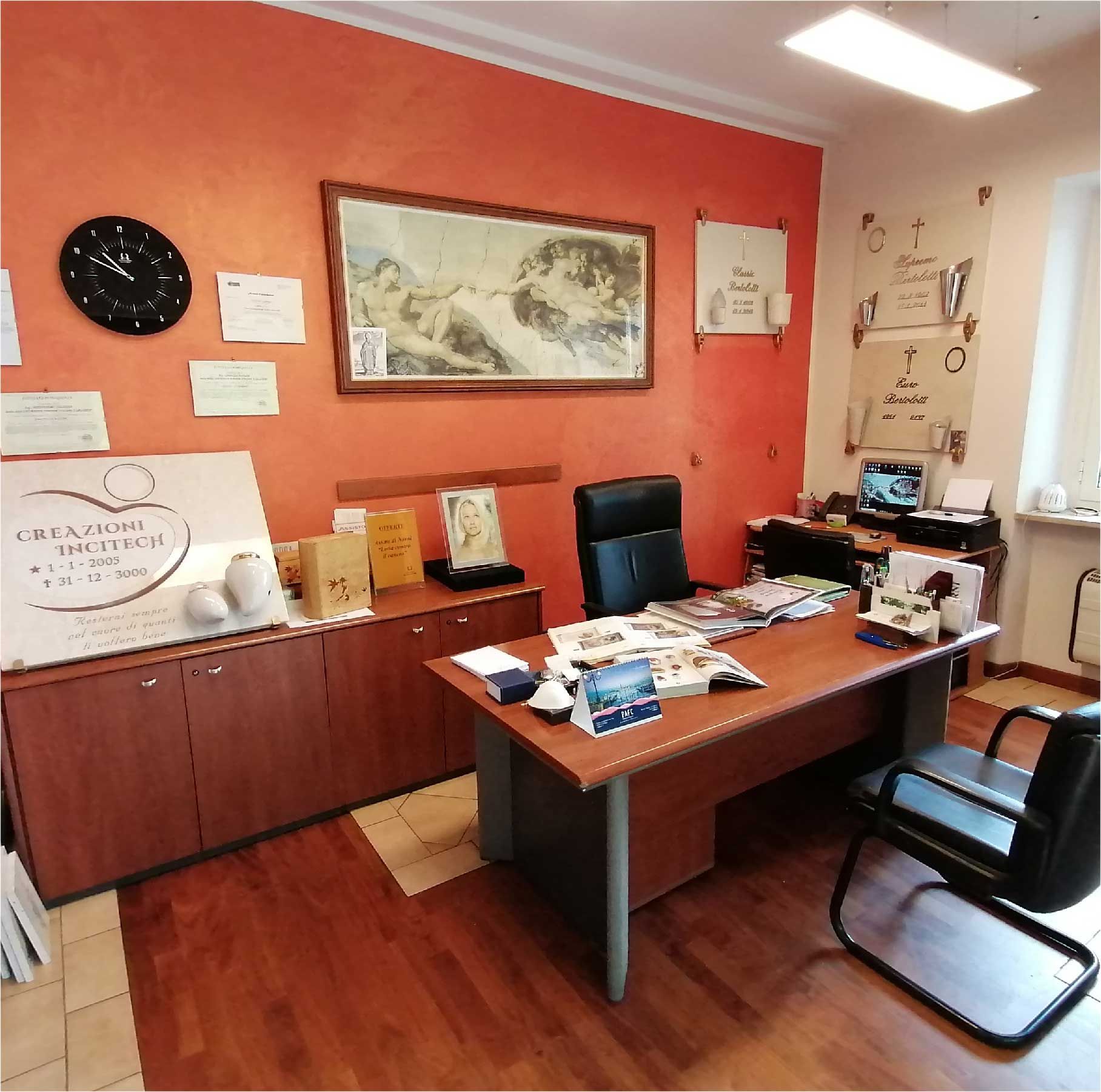 Piccolo ufficio con scrivania in legno, sedie, quadri alle pareti, orologio e fogli di carta su una parete arancione.