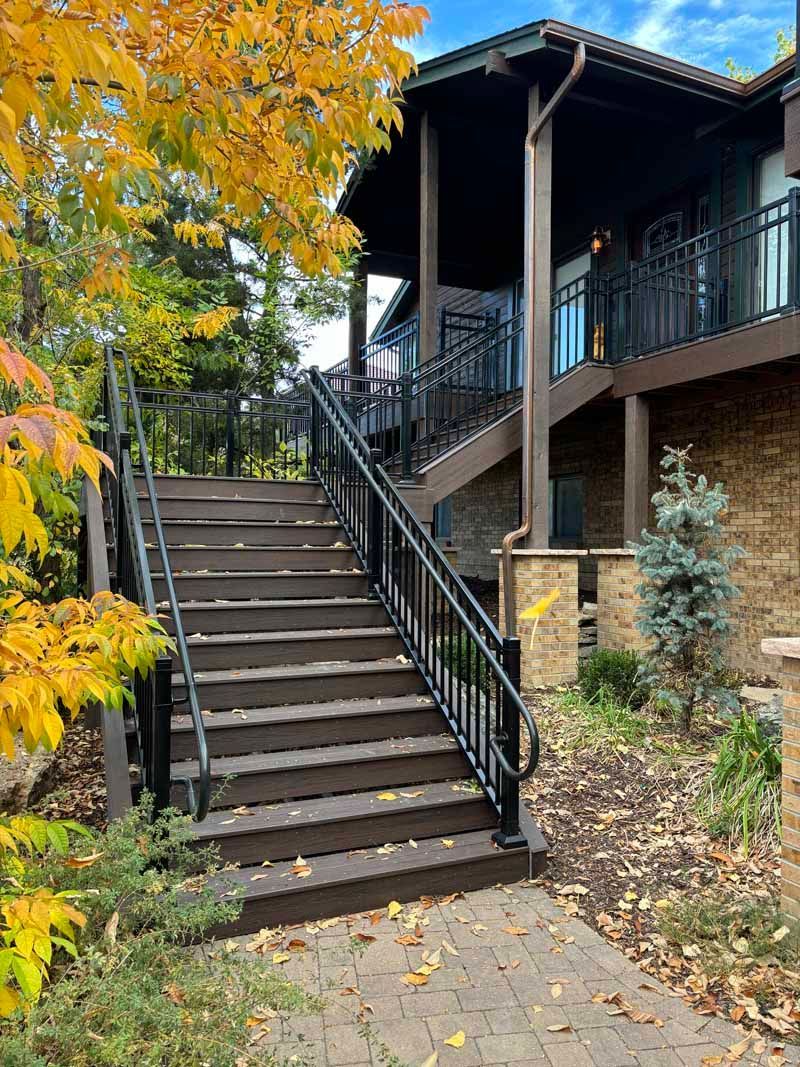 A set of outdoor wooden stairs with black railings leads up to a stone house deck surrounded by autumn trees.