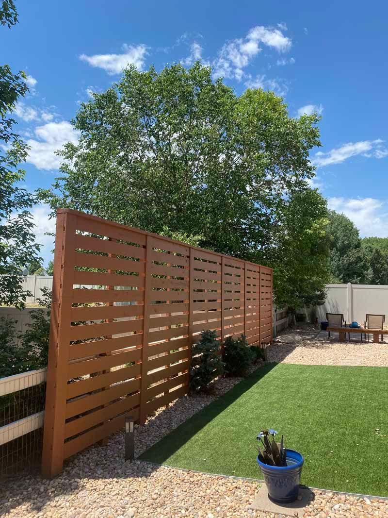 A tall, slatted wood privacy fence stands in a backyard with gravel mulch, a small patch of grass, and a blue pot.