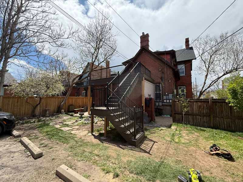 A backyard showing a multi-story brick house with a newly constructed wooden deck and staircase leading to a second floor.