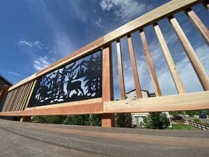 A wooden deck railing features a black metal panel cutout depicting a deer in a forest setting against a blue sky.