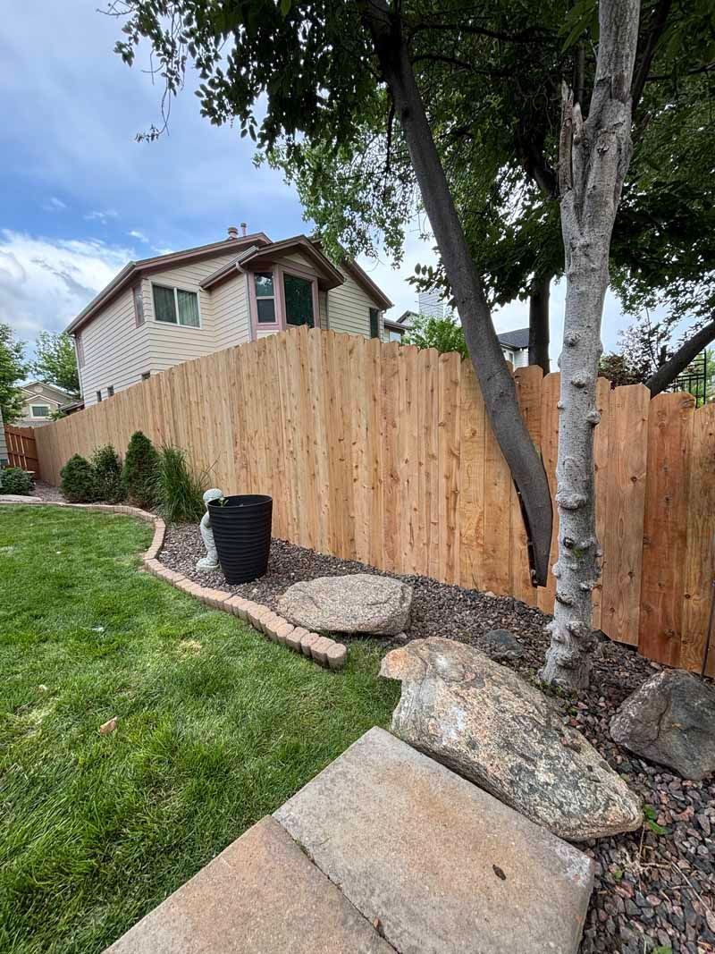 A sunny backyard featuring a new cedar wood fence, stone landscaping, large rocks, and trees next to a house.
