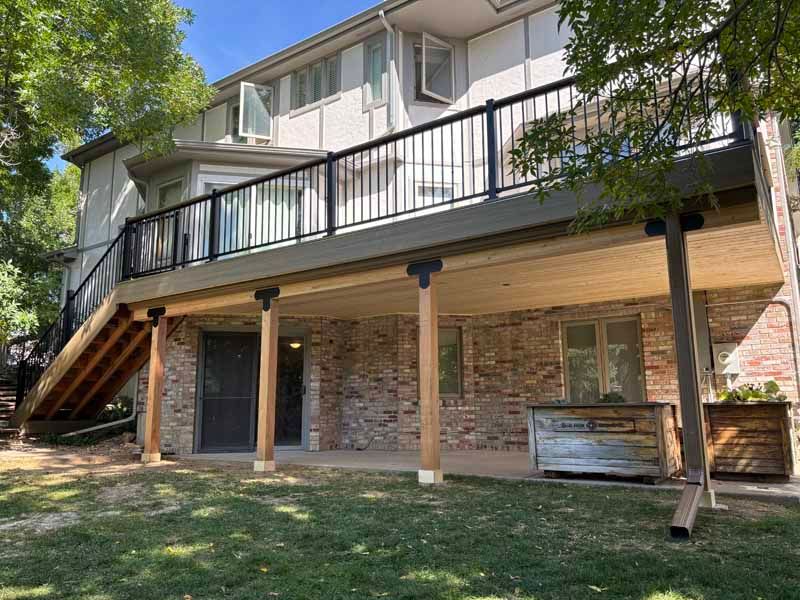A multi-level wood deck with black railings extends from the back of a two-story brick home, over a concrete patio.