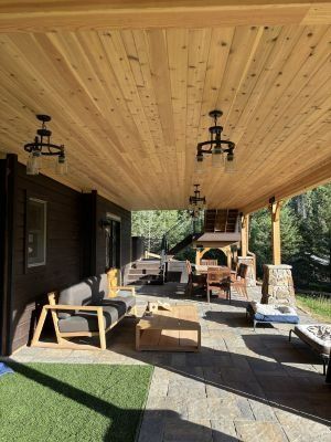 A covered outdoor patio featuring a wooden ceiling, a gray sofa, a wooden coffee table, and dining furniture on stone.