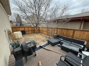 A patio with a grill and dark wicker furniture, leading to a small, fenced-in backyard with trees under a cloudy sky.