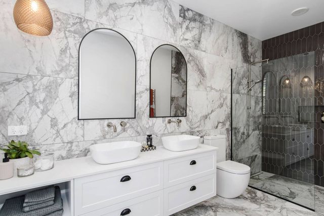 Modern bathroom with marble walls, double vanity, arched mirrors, and glass shower enclosure