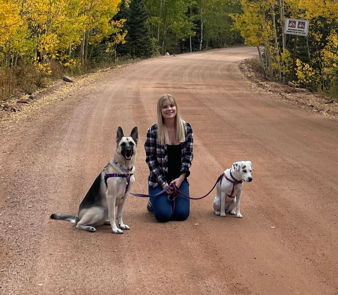 Smiling young woman posing on a dirt road with her two rescue dogs surrounded by autumn foliage