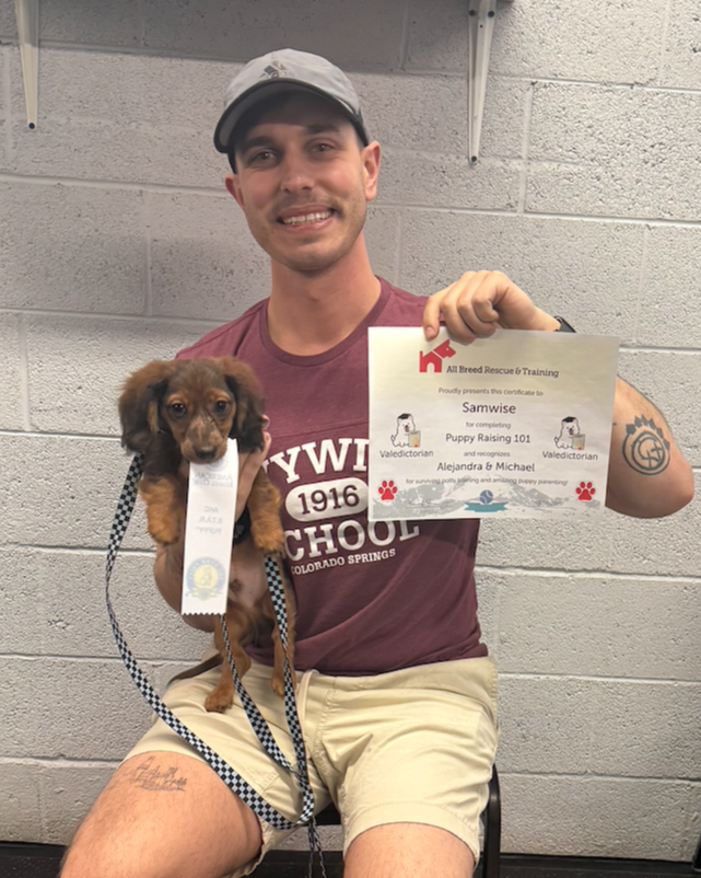 Happy puppy graduate holds up his grad certificate, while his puppy holds his AKC STAR Puppy ribbon