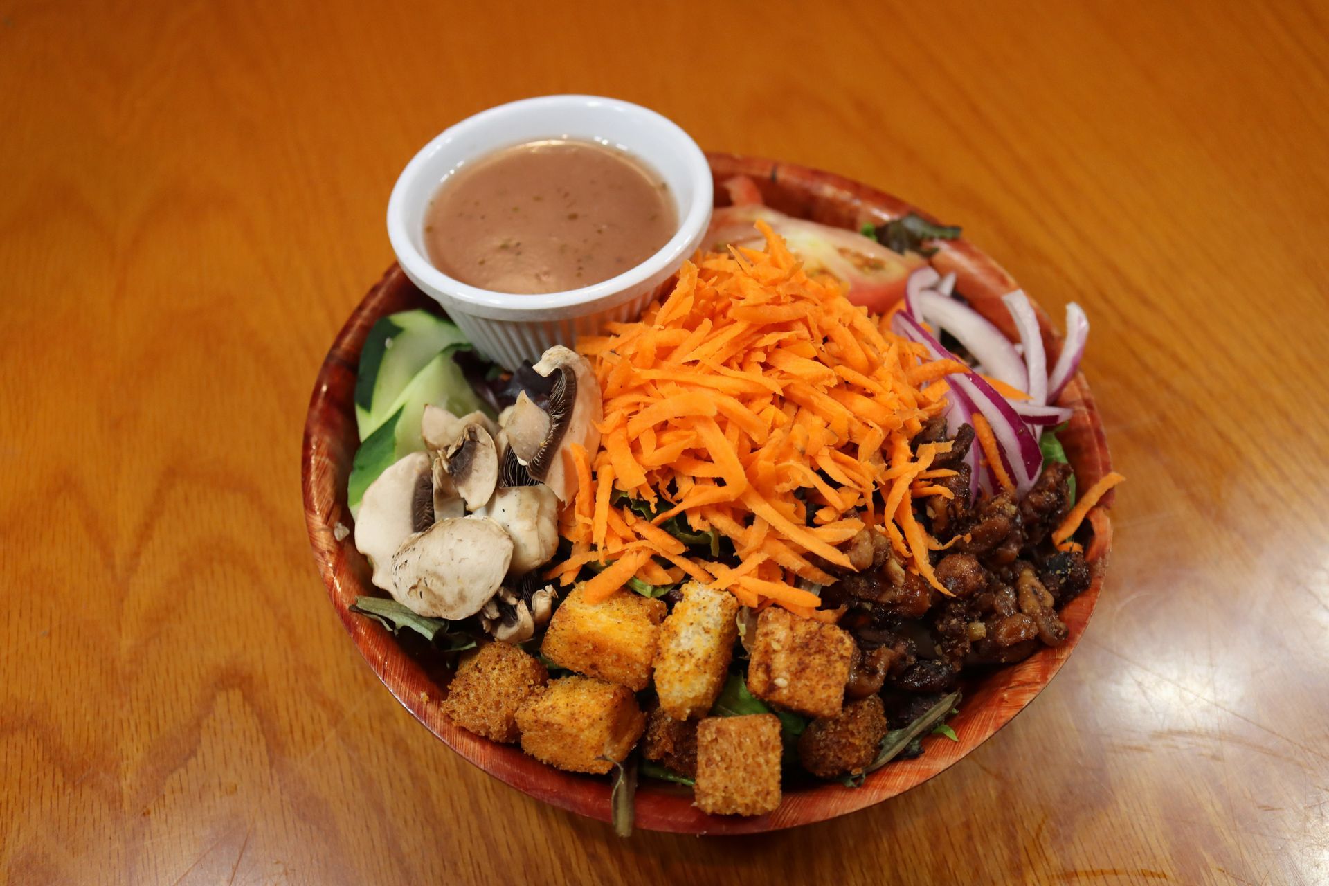 A salad with carrots , cucumbers , mushrooms , onions and croutons is on a wooden table.
