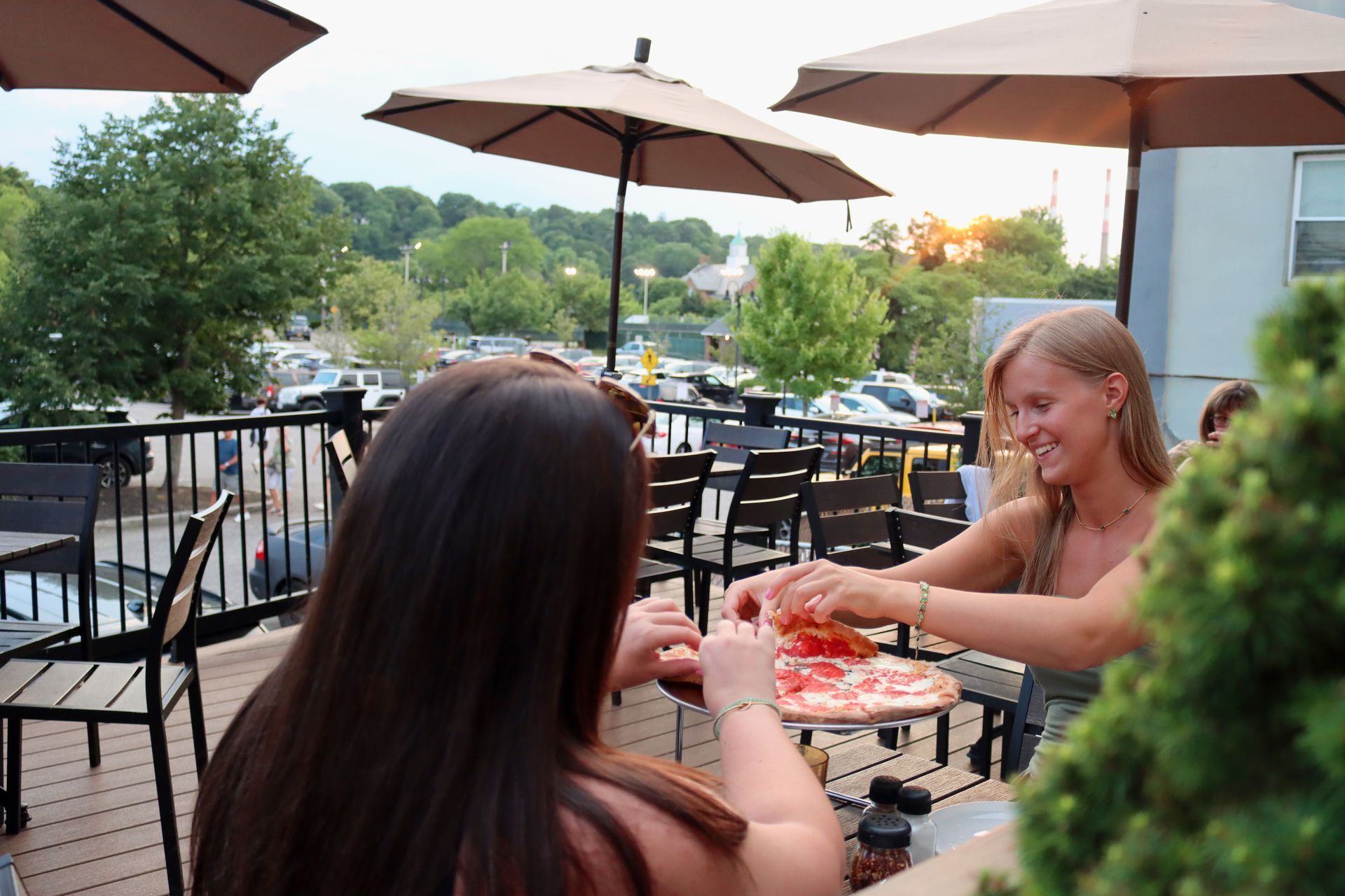 Two women are sitting at a table eating pizza under umbrellas.